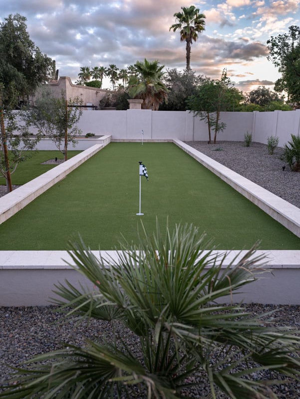Walled backyard putting green with tournament flags, travertine wall caps, and desert landscaping at sunset in Scottsdale AZ