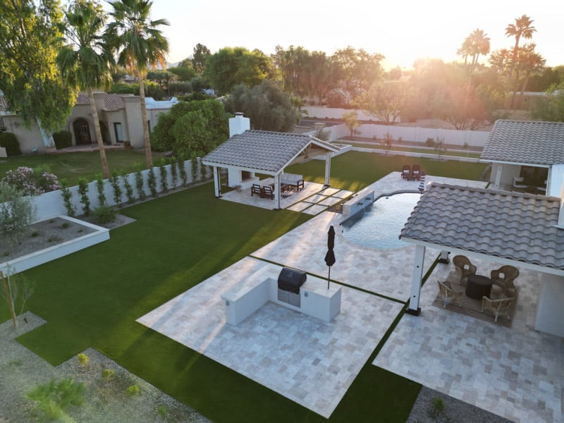Drone view of a complete Scottsdale backyard remodel with pool, twin pavilions, outdoor BBQ island, and artificial turf lawn at sunset